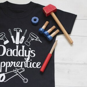 May include: A black t-shirt with white text that reads "Daddy's Apprentice". The shirt features a design of various tools, including a hammer, screwdriver, saw, and drill. The shirt is lying on a white wooden surface with toy tools scattered around it.
