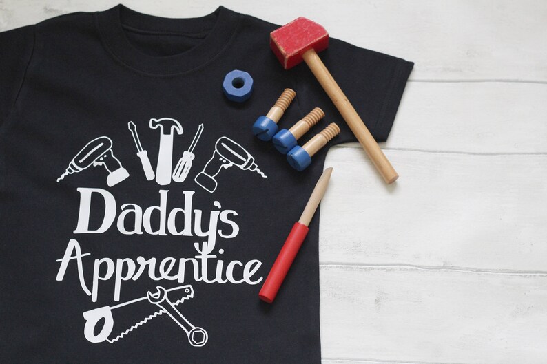 May include: A black t-shirt with white text that reads "Daddy's Apprentice". The shirt features a design of various tools, including a hammer, screwdriver, saw, and drill. The shirt is lying on a white wooden surface with toy tools scattered around it.