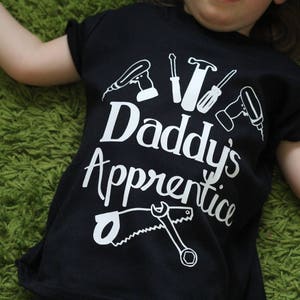May include: A black t-shirt with white text that says "Daddy's Apprentice" and a graphic of tools, including a saw, hammer, screwdriver, and drill.