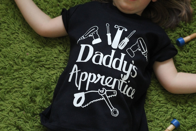 May include: A black t-shirt with white text that says "Daddy's Apprentice" and a graphic of tools, including a saw, hammer, screwdriver, and drill.
