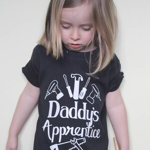 May include: A black t-shirt with a white design of tools and the text "Daddy's Apprentice". A young child is wearing the shirt and looking down.