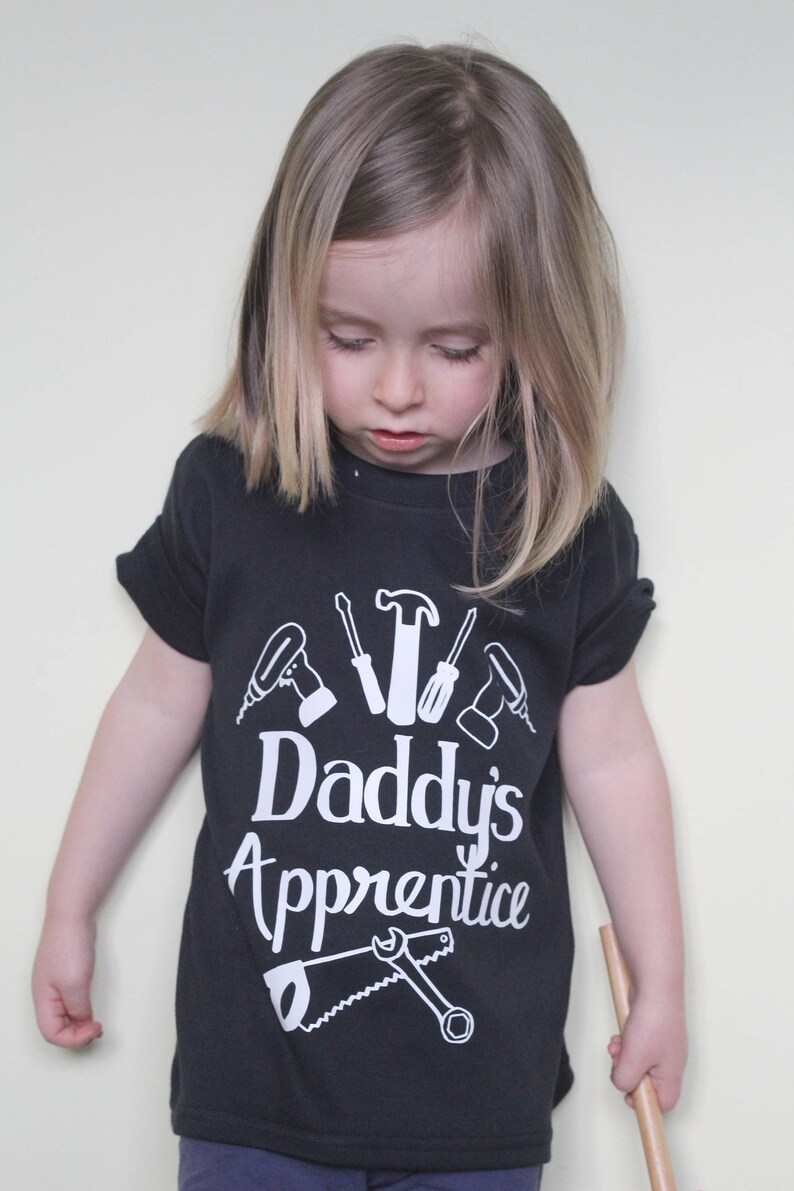 May include: A black t-shirt with a white design of tools and the text "Daddy's Apprentice". A young child is wearing the shirt and looking down.
