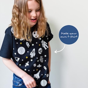 May include: A young person wearing a black t-shirt with a white space-themed design. The shirt features planets, stars, rockets, and an astronaut. The text "Make your own t-shirt" is written on a blue circle in the background.