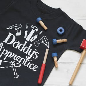 May include: Black t-shirt with white text that reads "Daddy's Apprentice" and a graphic of tools. The shirt is surrounded by toy tools, including a hammer, a saw, a screwdriver, and a wrench.