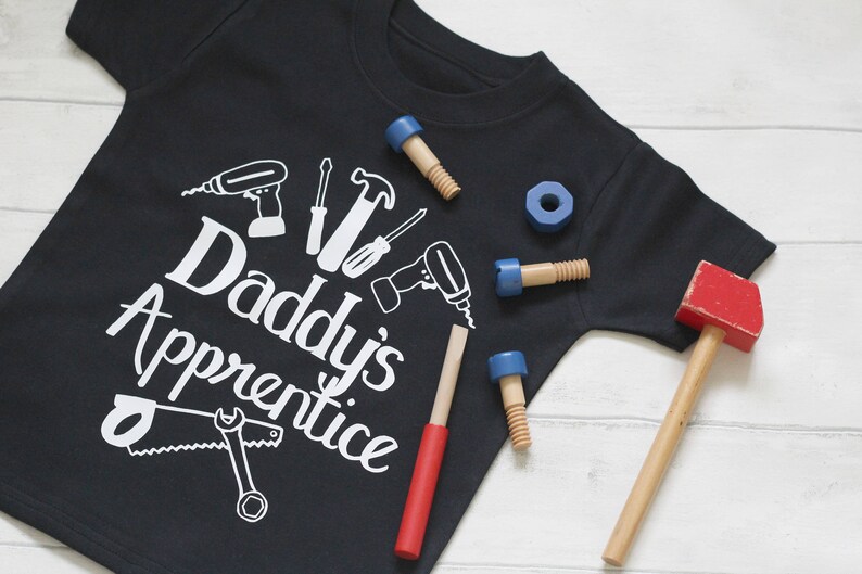 May include: Black t-shirt with white text that reads "Daddy's Apprentice" and a graphic of tools. The shirt is surrounded by toy tools, including a hammer, a saw, a screwdriver, and a wrench.