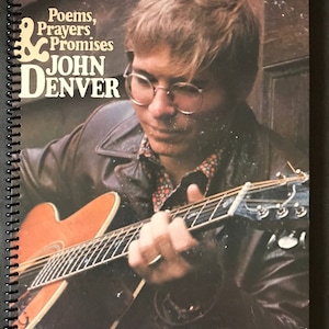 May include: A spiral-bound book cover featuring John Denver playing a guitar. The cover displays the text "Poems, Prayers & Promises" and "John Denver" in a vintage font. The image has a warm, nostalgic feel.