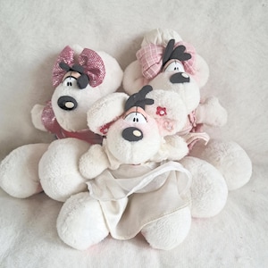 Op de afbeelding: Drie witte pluche knuffels met zwarte neuzen en donkere haarlokken. De linker draagt een roze strik met lovertjes en een topje. De achterste rechter heeft een roze geruite strik. De voorste draagt een witte jurk met een klein rood bloemdetail.