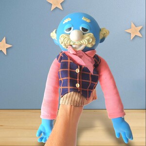 Op de afbeelding: Een blauwgezichtige handpop met een witte neus, snor en geel haar. De pop draagt een roze shirt, een geruit vest en een roze sjaal. De blauwe handen van de pop zijn zichtbaar. De achtergrond is een lichtblauwe muur met houten sterren.