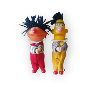 Op de afbeelding: Twee vintage Sesamstraat vingerpoppen. De pop links heeft zwart haar, een rood-wit gestreept shirt en een rode broek. De pop rechts heeft geel haar, een geel-wit gestreept shirt en een gele broek.