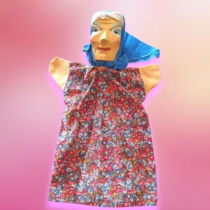 Op de afbeelding: Een vintage pop met een gesneden houten gezicht met geschilderde details, een blauwe sjaal en een jurk met bloemenprint. De armen van de pop zijn gemaakt van beige stof. De jurk heeft een patroon van rode, blauwe en gele bloemen.