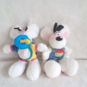 May include: Two plush mouse toys with white fur and large ears. The left mouse wears a blue bib with red dots, a yellow shirt, and a blue pacifier. The right mouse wears blue denim-style overalls with a red pocket detail and has black hair.