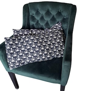 Op de afbeelding: Een donkergroene fluwelen fauteuil met een gecapitonneerde rugleuning. Een rechthoekig kussen met een herhalend patroon van zwarte kattengezichten op een witte achtergrond ligt op de stoel.