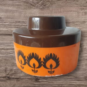 Op de afbeelding: Een bruine en oranje plastic container met een deksel. De container heeft een ontwerp met drie zwarte vlammen aan de voorkant.