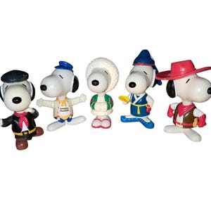 May include: Five Snoopy figurines in various outfits. One wears a black beret and jacket, another a blue cap and patterned jacket, a third a white winter hat and green coat, a fourth a blue hat and kimono, and the last a red cowboy hat and vest.