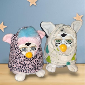 Puede incluir: Dos juguetes Furby vintage. Uno es rosa con pelo azul y una capucha con lunares negros y rosas. El otro es blanco con una capucha con lunares blancos y negros y detalles de pera verde. Ambos tienen picos amarillos y ojos grises.