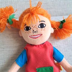 Puede incluir: Mu&ntilde;eca de peluche con coletas de hilo naranja atadas con cintas verdes. La mu&ntilde;eca tiene una cara sonriente con rasgos bordados, incluidas pecas, y viste un vestido rojo con mangas azules. Juguete infantil.