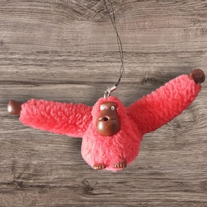 Peut inclure: Un porte-clés en forme de singe en peluche rose vif avec des yeux bruns et un nez brun. Le singe est suspendu à une ficelle noire.