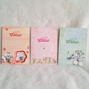 May include: Three rectangular paper notepads featuring the Diddl logo. The left pad is peach with a flower-shaped cutout and mouse illustrations. The center pad is pink with a grid pattern and small mouse graphics. The right pad is light blue with musical notes and a mouse playing a flute.
