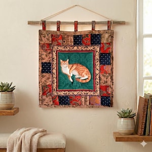 Op de afbeelding: Een vierkante stoffen wandhanger met een centraal paneel met een oranje cyperse kat op een groene achtergrond, omringd door een patchworkrand van rode, blauwe en beige bloemen- en stippenpatronen, opgehangen aan een houten stok met een touw.