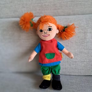 Puede incluir: Mu&ntilde;eca de peluche con coletas naranjas, pecas y una cara sonriente. La mu&ntilde;eca viste una camisa azul, un chaleco rojo con un bolsillo verde, pantalones verdes, calcetines desparejados y zapatos negros. Juguete infantil.