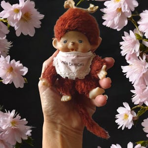 Op de afbeelding: Een bruine pluche apenpop met een roze slab waarop "Mon Chichi" staat, wordt in een hand vastgehouden. De aap draagt ​​een bruine bontmuts met geweien. De achtergrond is een wazig beeld van roze bloemen.