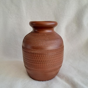 Op de afbeelding: Een vaas van terracotta-kleurige klei met een rond lichaam en een brede, gladde rand. Het buitenoppervlak is versierd met horizontale banden van ingewikkelde, herhalende geometrische patronen, waaronder kruisarceringen en kleine cirkelvormige inkepingen.