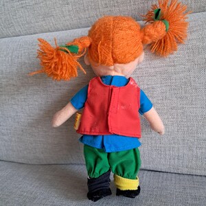 Puede incluir: Mu&ntilde;eca de peluche con coletas de hilo naranja atadas con cintas verdes. La mu&ntilde;eca viste una camisa azul, un chaleco rojo, pantalones verdes y calcetines negros y amarillos. Juguete infantil.