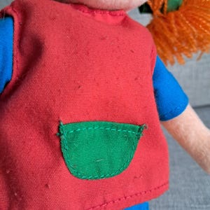 Puede incluir: Primer plano de una mu&ntilde;eca que lleva un chaleco rojo con un bolsillo verde sobre una camisa azul. La mu&ntilde;eca tiene el pelo naranja. El chaleco tiene algunas peque&ntilde;as imperfecciones.