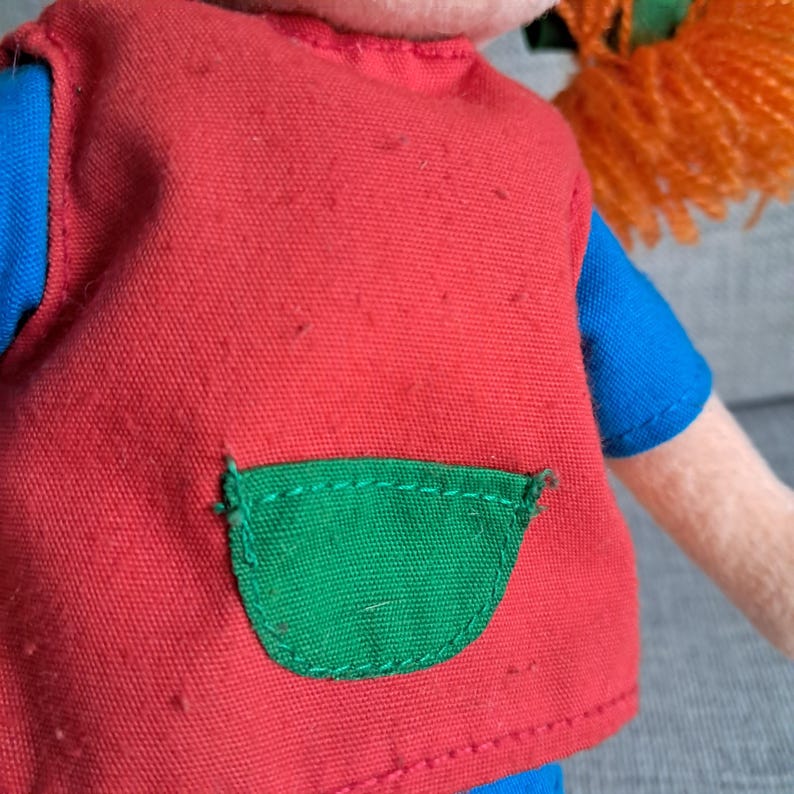Puede incluir: Primer plano de una mu&ntilde;eca que lleva un chaleco rojo con un bolsillo verde sobre una camisa azul. La mu&ntilde;eca tiene el pelo naranja. El chaleco tiene algunas peque&ntilde;as imperfecciones.
