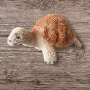 Könnte beinhalten: Ein Plüschtier-Schildkröte mit einer braunen Schale und einem hellbraunen Körper. Die Schildkröte hat einen kleinen, runden Kopf mit schwarzen Augen und einer kleinen, rosafarbenen Nase.