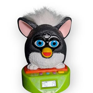 Op de afbeelding: Een vintage Furby-wekker. De Furby heeft een zwarte body met witte accenten, blauwe ogen en een gele snavel. De klokbasis is groen met een oranje bovenkant, met knoppen en een digitale display met de tekst "10:19".