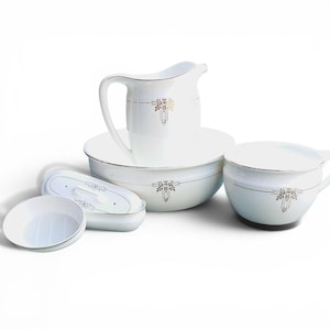 May include: A white porcelain wash set with gold trim and floral detailing. The set comprises a jug, basin, covered dish, soap dish, and small bowl. The items are arranged on a white background.