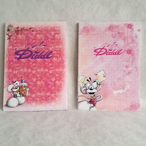 Op de afbeelding: Twee rechthoekige papieren notitieblokken met het Diddl-logo in paars en roze. Het linkerblok heeft een donkerroze bloemenpatroon met een stripmuis die een verjaardagstaart vasthoudt. Het rechterblok heeft een lichtroze raster met een stripmuis die brieven vasthoudt.
