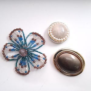 Op de afbeelding: Een verzameling van drie broches: een bloemvormige broche gemaakt van blauwe, bruine en doorzichtige kralen; een ronde broche met een crèmekleurige steen in het midden omringd door kleine witte parels; en een ovale broche met een donker metalen centrum.