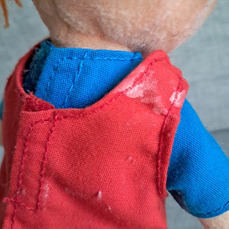 Puede incluir: Primer plano del hombro y la parte superior del torso de una mu&ntilde;eca. La mu&ntilde;eca lleva un chaleco rojo brillante sobre una camisa azul. El chaleco tiene marcas de pintura blanca. La piel de la mu&ntilde;eca es de color melocot&oacute;n claro y el pelo es naranja.