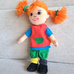 Puede incluir: Mu&ntilde;eca de peluche con coletas naranjas brillantes, pecas y una cara sonriente. Viste un chaleco rojo sobre una camiseta azul, pantalones verdes y calcetines desparejados. Zapatos negros. Un juguete infantil cl&aacute;sico.