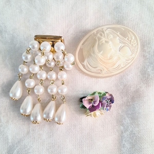 Op de afbeelding: Een goudkleurige broche met trapsgewijze strengen witte kunstparels, een ovale, crèmekleurige cameebroche met een reliëf van twee gezichten, en een kleine ring versierd met een trosje beschilderde keramische viooltjes in paars en groen.