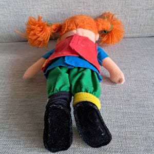 Puede incluir: Mu&ntilde;eca de peluche con coletas de hilo naranja brillante, vestida con una camisa azul, un chaleco rojo y pantalones verdes. La mu&ntilde;eca tiene zapatos negros y calcetines negros y amarillos. Est&aacute; sobre una superficie gris.