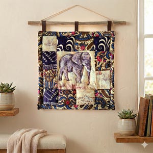 Op de afbeelding: Een vierkante stoffen wandhanger met een centraal paneel met een grijze olifantillustratie, omringd door een patchworkrand van donkerblauwe, bruine en crèmekleurige bloemen- en geometrische patronen, opgehangen aan een houten stok.