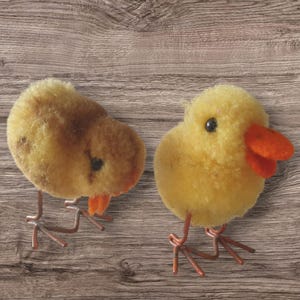 Op de afbeelding: Twee gele, pluche kuikenfiguren met oranje snavels en zwarte ogen. De kuikens staan op draadpoten tegen een bruine houten achtergrond.