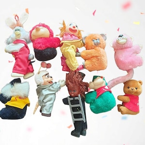 May include: A collection of vintage plush toys, including a white rabbit in pink overalls, a clown in yellow, and several bears. The toys are in various colors, such as pink, orange, and brown, and are arranged against a white background.