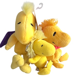 Op de afbeelding: Een cluster van gele pluche speelgoed met het personage Woodstock uit de strip Peanuts. Eén figuur heeft een paarse vleugel. Het speelgoed heeft zwarte ogen en is gemaakt van zachte, gele stof. Een klein label is bevestigd.