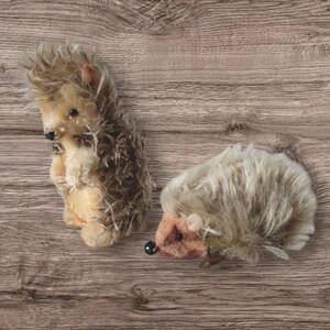 Puede incluir: Dos juguetes de peluche de erizo marrón y gris con ojos y narices negros. Los erizos están sobre un fondo de madera.