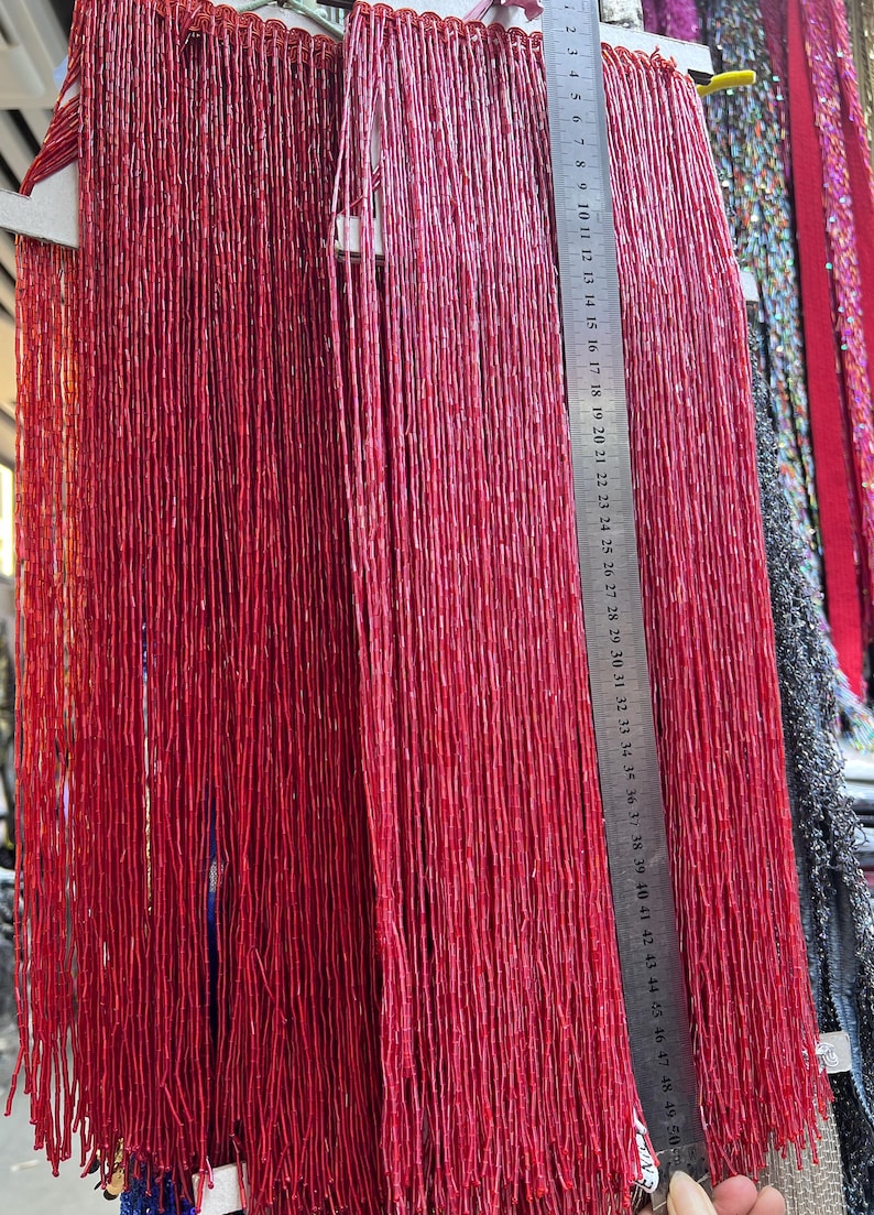 May include: Close-up of red beaded strands hanging vertically. A metal ruler is placed next to the strands, showing measurements up to 50 cm. The beads appear to be small and cylindrical, creating a textured, vibrant display.