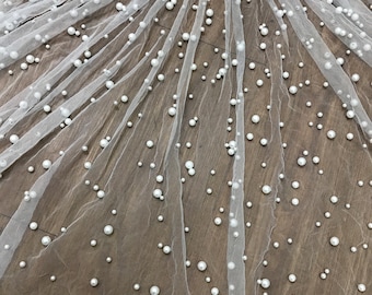 May include: White tulle fabric with white pearl embellishments. The fabric is laid out on a wooden surface.
