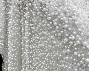 May include: Close-up of a white fabric heavily embellished with numerous white pearls of varying sizes. The pearls are densely packed, creating a textured, shimmering surface. Small silver sequins are interspersed among the pearls, adding extra sparkle.