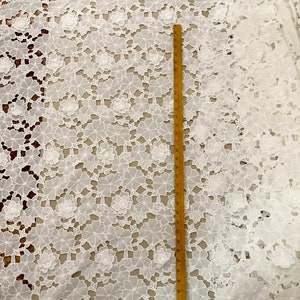 May include: White cotton lace fabric with a floral pattern. The fabric is shown with a ruler measuring 30 inches.