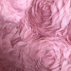 以下が含まれることがあります： ピンクの生地のクローズアップで、3Dのバラ模様が施されています。生地は柔らかく繊細な素材でできており、上品な光沢があります。