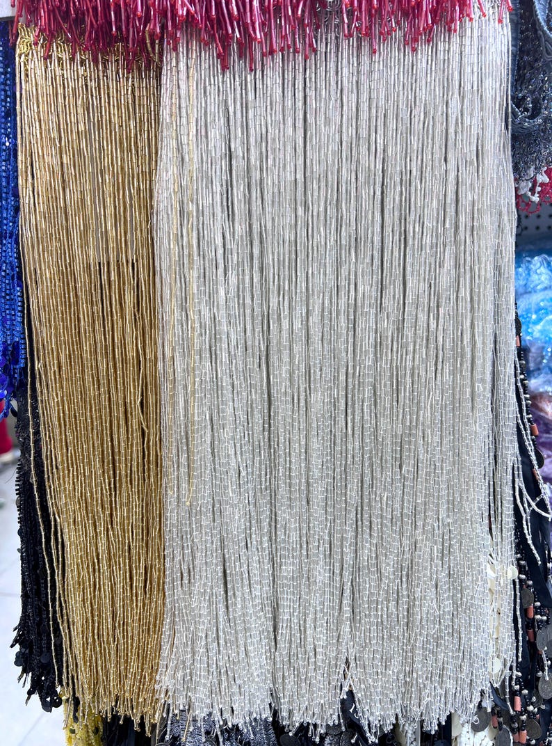 May include: Close-up of beaded fringe in gold, silver, and red. The strands are tightly packed, creating a textured surface. The beads appear to be small and cylindrical, giving the fringe a shimmering effect. Ideal for embellishments.