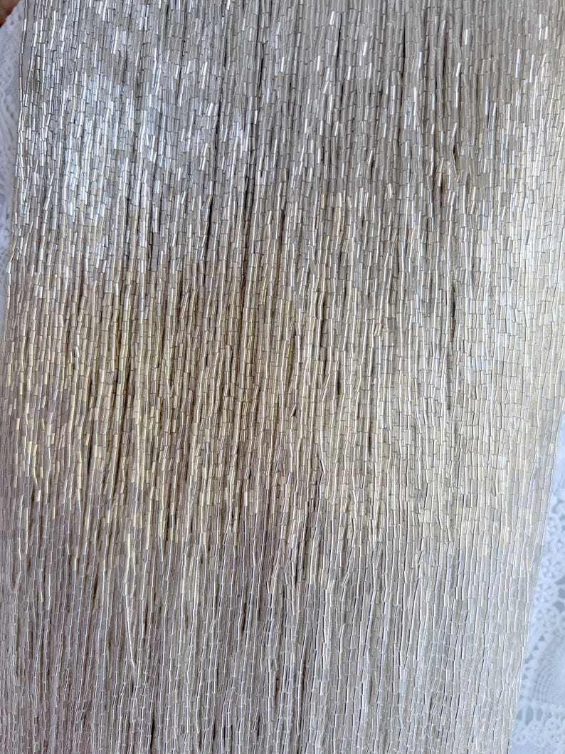 May include: Close-up of a silver beaded fabric. The beads are small, cylindrical, and densely packed, creating a shimmering, textured surface. The beads are arranged in vertical rows, giving the fabric a draped appearance. The background is a soft, off-white color.